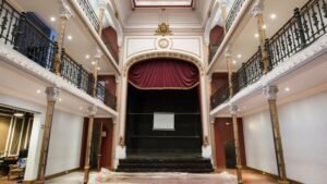 Interior del Teatro Huemul, al fondo se encuentra el escenario con fondo negro y cortinas rojas, encima de lo anterior cuelgan dos logos, el del teatro y el de la caja de ahorro. A los costados se aprecian las galerías blancas con rejas de fierro. En último lugar está el patio, frente al escenario, sin ningún accesorio.