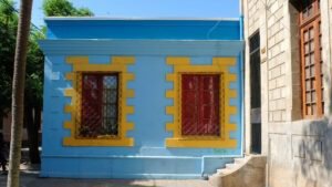 Imagen en colores del costado de una casa de la Población Huemul I. La casa es de un piso y de construcción de hormigón propia del barrio. La casa está pintada en colores celeste y amarillo y las ventanas son rojas. A la derecha se ve parte del costado del edificio de la Caja de Ahorros.