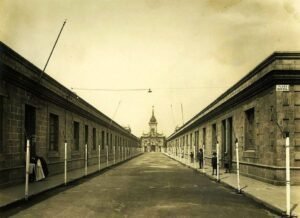 Fotografía en color sepia de Calle Los Algarrobos, en ambos costados se encuentran casas pareadas de un piso y gente caminando por las veredas sin árboles. Al final de la avenida se encuentra la iglesia Santa Lucrecia.