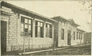 Imagen en blanco y negro del frontis del edificio de la Escuela de Mujeres. Es una construcción de un piso, con paredes de hormigón y ventanas a lo largo de todo el frontis y una puerta principal. La calle es de tierra y se ven árboles pequeños y sin hojas en la vereda.