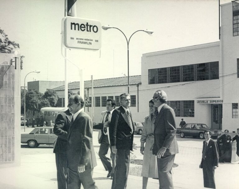 Fotografía en blanco y negro en donde se observan en primer plano varios hombres y una mujer con tenidas formales, sonrientes y hablando en las afueras de la estación. En segundo plano, se observa a otras personas mirando la escena y en la parte superior de la fotografía un cartel que dice: Metro, acceso estación, Franklin. En tercer plano, se ven autos de los años 70 y la Escuela Superior Hermanos Matte.