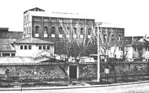 Fotografía en blanco y negro del edificio de la ex Refinería de Azúcar. El plano es general y se ve gran parte del edificio el cual posee una muralla alta alrededor, luego se observan construcciones de uno y dos pisos, y finalmente, más hacia el fondo y destacando, se observa el edificio más alto con grandes ventanas y techos muy altos.