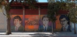 Fotografía del memorial por los ejecutados políticos del Barrio Huemul. Es un mural en el que se observan los rostros de 3 ejecutados políticos junto a una leyenda escrita sobre fondo de colores cálidos. La leyenda dice: Aunque los pasos toquen mil años este sitio no borrarán la sangre de los que aquí cayeron.