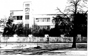 Imagen en blanco y negro del frontis del edificio de la escuela. En primer plano, se observan árboles medianos, la calle, la vereda y una persona que camina. En segundo plano, se observa el edificio, el cual posee tres pisos y una torre con un reloj a la izquierda.