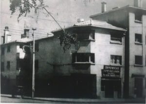 Imagen en blanco y negro del frontis del complejo habitacional Huemul III compuesto por un edificio de cuatro pisos en el extremo izquierdo y dos de tres pisos en el centro de la fotografía. En la esquina inferior izquierda se observa un local de comida rápida llamado “Sandwich Americanos”.