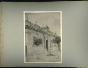 Imagen en blanco y negro del frontis del edificio de la Gota de Leche. La fotografía está puesta sobre un papel gris de álbum de fotos. El frontis es de color claro, la construcción posee un piso y se ven algunos árboles pequeños en la vereda.