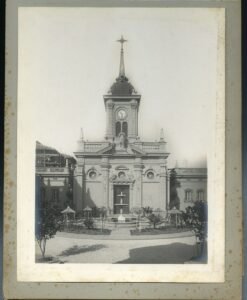 Imagen en blanco y negro de la Iglesia Santa Lucrecia. En el centro se observa la cúpula de la parroquia. A los costados están el ala izquierda y derecha de la parroquia con dos ventanas cada una. En frente de la edificación se encuentra una pileta de mármol. La fotografía está puesta sobre papel gris de álbum de fotos.