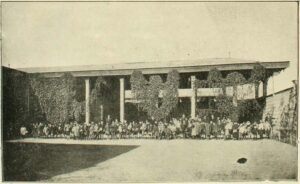 Imagen en blanco y negro de un plano general del patio interior de la Escuela de Hombres del barrio. Al centro se observan gran cantidad de niños dispuestos en una hilera horizontal de izquierda a derecha. Entre medio se observan algunos adultos y adultas. En la parte superior se ve el techo de un galpón y enredaderas que cuelgan de este. En la parte inferior se ve una cancha de pasto.