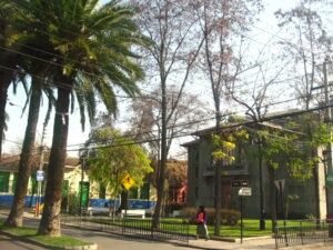 Fotografía en colores de la Plaza Huemul sector Teatro Huemul. Se observa en primer plano dos palmeras, la calle asfaltada y una reja verde que rodea la plaza. Luego, una persona que camina por la vereda. En tercer plano, se observan los árboles y pasto de la plaza y el Teatro Huemul y ex Escuela de Hombres al fondo.