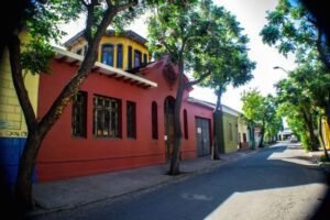 Imagen en colores del frontis del edificio de la Biblioteca desde una esquina. El edificio es color rojo oscuro y posee un torreón es amarillo. Se ve la calle vacía y muchos árboles verdes en las veredas.