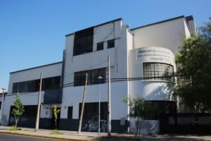 Imagen en colores del frontis del edificio de la escuela, vista lateral izquierda. En primer plano, se observa la calle, la vereda y algunos árboles. En segundo plano, se observa el edificio, el cual posee dos pisos a la izquierda, y tres pisos en el centro y derecha. La esquina es redondeada y se lee la leyenda: Escuela Hermanos Matte, Sociedad de Instrucción Primaria, Año 1935.