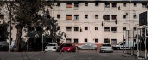 Fotografía actual de un edificio de departamentos desde el interior de la Población Huemul II. La construcción tiene 4 pisos y las paredes son de color beige. Se observa una cancha y estacionamientos.