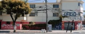 Fotografía actual de la fachada lateral de uno de los edificios de la Población Huemul III. Sus paredes tienen color rojo en el primer piso y beige en el segundo y tercer piso. Las paredes están rayadas con grafitis. Se observa un árbol a la izquierda del edificio.