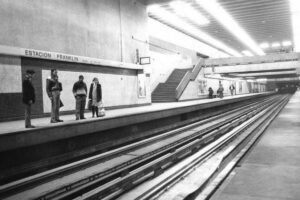 Fotografía en blanco y negro del andén de la estación de metro Franklin, correspondiente a la línea 2. Se observa un grupo de personas que esperan la llegada del vagón y otras caminando al fondo de este. En el fondo se observa la escalera que da al cambio de andén y a la salida de la estación.