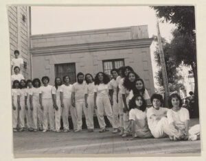 El grupo de aproximadamente 20 hombres y mujeres se encuentra en el exterior izquierdo del Teatro Huemul. Captura en blanco y negro.