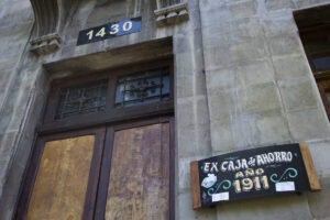 Imagen que muestra un detalle del frontis del edificio, se ve la parte superior de la puerta de madera y al lado derecho de la imagen se observa una placa de madera con fondo negro y letras blancas y amarillas escritas con la tipografía tradicional de los carteles de locomoción colectiva chilenos, en las que se lee: Ex Caja de Ahorro, año 1911.