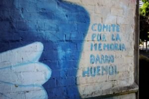 Al costado derecho del memorial, en la muralla que da vuelta la esquina, se observa una ala de paloma y en fondo gris se ven letras celestes que dicen: Comité por la memoria Barrio Huemul.