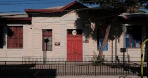 Frontis del edificio de la ex Escuela de Mujeres. Se mantiene la construcción de un piso, con paredes de hormigón, ventanas a lo largo del frontis y una puerta principal. La calle es de cemento y se ven árboles en la vereda. Al lado de la puerta principal se observan placas con el nombre del establecimiento.