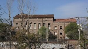 Fotografía del lado poniente del edificio de la ex Refinería de Azúcar. El plano es general y se ve gran parte del edificio el cual posee una muralla alta alrededor, y tras esta un edificio alto con grandes ventanas, algunas están rotas.