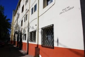 Fotografía en colores de la vista lateral e interior de uno de los edificios de la población Huemul II en la actualidad. Se observan 4 pisos, ventanas pequeñas en cada piso con rejas. El edificio está pintado con una franja roja en la parte inferior y el resto color beige. En la pared se lee: pasaje profesor Juan A. Villagrán.