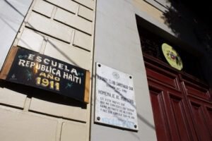 Imagen en color de un detalle del frontis del edificio de la Escuela de Mujeres. De izquierda a derecha se observan distintas placas. La primera es de madera, fondo negro y letras blancas y amarillas, la cual está realizada en la tipografía y estilo de los carteles tradicionales de la locomoción colectiva chilena. La segunda placa, pertenece al Rotary Club Santiago Sur y se lee: Rotary Club Santiago Sur, Semana del Niño, En este nuevo día que me concedes oh señor, dame mi parte de alegría y haz que consiga ser mejor. 1960. Gabriela Mistral. La tercera placa, es la tradicional placa de las escuelas públicas chilenas, que contiene el escudo nacional y se lee Escuela Pública Chile.