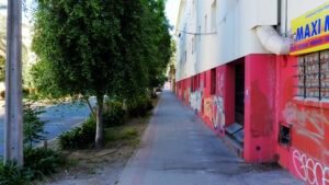 Imagen en colores del costado de uno de los edificios de la Población Huemul III en la actualidad. Al lado izquierdo se encuentra la calle y árboles, en el centro la vereda y a la derecha está el edificio. La parte inferior está pintada roja y la superior es beige, se ven muchos rayados en la pared y un cartel de negocio en el costado derecho de la imagen.