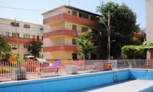 Fotografía en colores de la piscina en el interior de la población Huemul II en la actualidad. Se observan 3 edificios de 4 pisos, con pasillos laterales y ventanas pequeñas en cada piso. El edificio está pintado con una franja roja en la parte inferior y barandas y el resto color beige. La piscina está vacía.