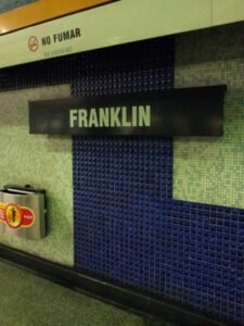 Cartel en color negro y letras blancas al interior del andén línea 2 de la estación Metro Franklin en la actualidad, se lee: Franklin. De fondo se observa una pared con mosaicos azules y grises. Sobre el cartel, en la parte superior, dice No Fumar.