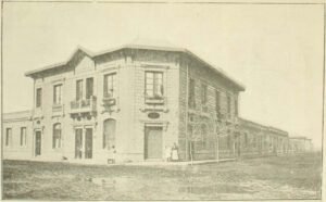 Edificio casa esquina de dos pisos en una intersección de dos calles grandes no asfaltadas. Mujeres y niños se encuentran frente a la edificación. Plantas cuelgan de los balcones. Se ven pozas de agua en la calle y árboles sin hojas en las veredas.