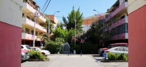 Imagen en colores de la entrada al complejo habitacional Huemul III. Se observan edificios de 4 pisos a los costados y un patio interior al centro de la fotografía, con plantas, arbustos, árboles, algunos autos y una gruta cerrada con una reja celeste.