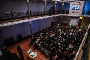 Interior del Teatro Huemul, se observa una persona que está dictando un seminario, enfrente de él, en el patio del teatro, una treintena de hombres y mujeres están sentados mirándolo a él. Las galerias que rodean el patio son de fierro negro.