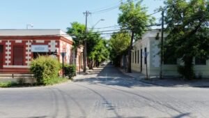 Imagen en colores de una calle en la Población Huemul I. En primer plano se ve la calle con asfalto y adoquines, luego se ven dos casas esquina, una de colores rojo y blanco, y otra de colores blanco invierno y blanco. Ambas en construcción de hormigón propia del barrio.