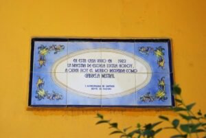 Imagen en colores de placa de cerámica ubicada en el frontis de la casa de Gabriela Mistral en la actualidad. La pared es amarilla y en el centro de la imagen se observa la plaza que dice: En esta casa vivió en 1922 la maestra de escuela Lucila Godoy, a quien hoy el mundo recuerda como Gabriela Mistral. Ilustre Municipalidad de Santiago, Departamento de Cultura.