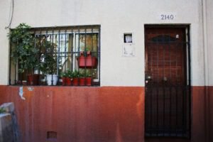 Fotografía en colores de la fachada de un departamento de la población Huemul II. A la izquierda se ve una ventana, con rejas negras y plantas en maceteros. A la derecha se ve una puerta de madera con una reja negra y un medidor de luz. La parte inferior está pintada de color rojo y la parte superior de color beige.