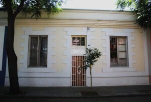 Imagen en colores de una casa en la Población Huemul I. La casa es de un piso y es de construcción de hormigón propia del barrio. Tiene dos ventanas a los costados y una puerta delgada y alta al centro. La casa está pintada de colores beige y blanco.