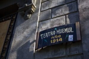 Cartel ubicado al costado de la puerta principal del Teatro Huemul. En él se escribió con tipografía cursiva y en colores brillantes sobre un fondo oscuro. Se lee “Teatro Huemul, año 1914”.