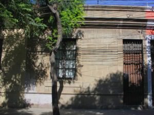 Imagen en colores de una casa en la Población Huemul I. La casa es de un piso y es de construcción de hormigón propia del barrio. Tiene una ventana al costado izquierdo y una puerta delgada y alta al costado derecho. La casa está sin pintar con el color original del hormigón.